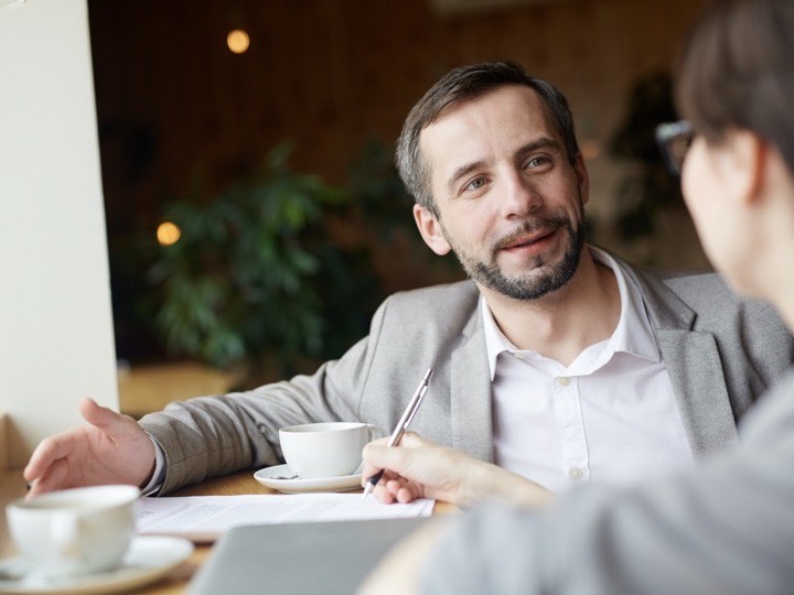 Dos personas conversando en una mesa de café con tazas y una libreta